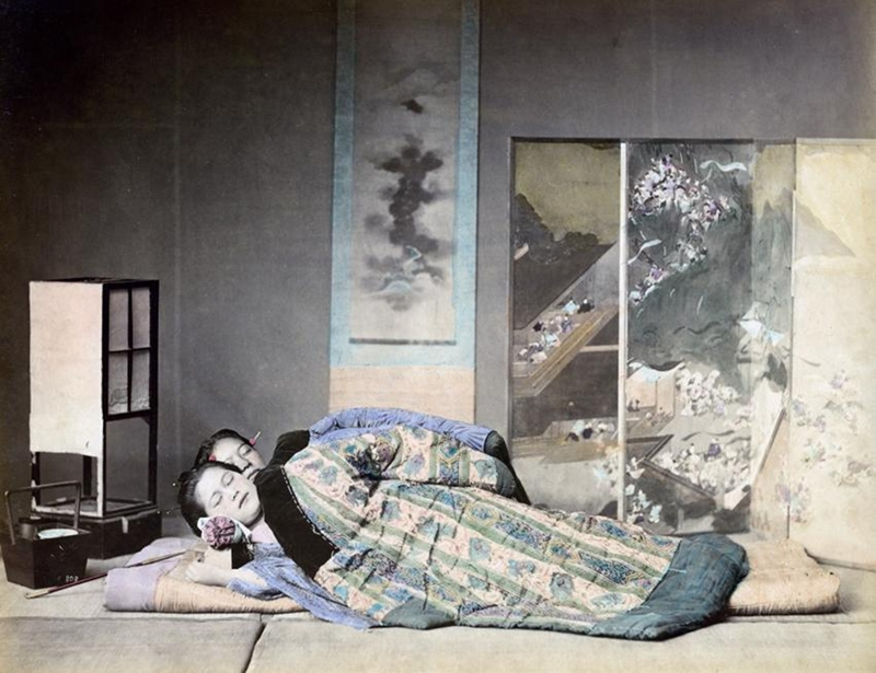 Two women sleeping under a padded coat / blanket in a room with painted screen, scrolled painting and paper lantern. The cushion is a small wooden box