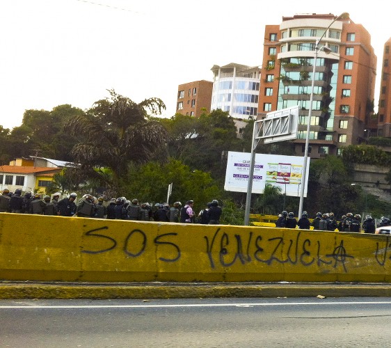 Grafiti de #SOSVenezuela en la autopista y detrás soldados de la Guardia Nacional Bolivariana mirando la manifestación abajo. Foto de Kira Kariakin.