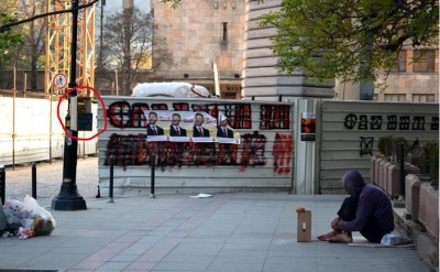 Scéna z ulic Skopje. V popředí krabice pro darování jídla a žebrající člověk. V pozadí volební plakáty a staveniště nové vládní budovy. Foto organizace Share food.