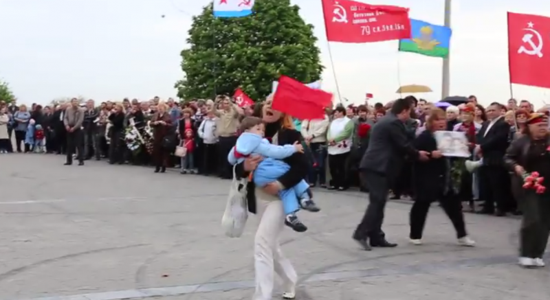 A woman carrying a child charges the stage in disgust, after the Governor speaks ill of the Red Army and ambiguously about Hitler. Kherson, Ukraine. May 9, 2014. YouTube.