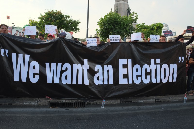 Протестующие против переворота, собравшиеся на площади перед Монументом Победы в Бангкоке, держат огромный плакат с надписью «Мы хотим Выборы»
