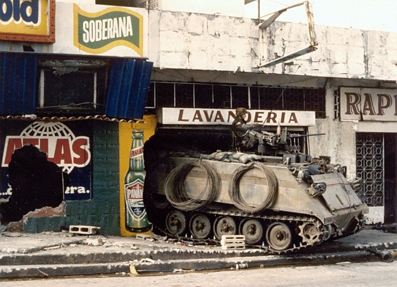 A U.S. Army M113 armored personnel carrier guards a street near the destroyed Panamanian Defense Force headquarters building during the second day of Operation Just Cause. Photo by J. Elliott, public domain.